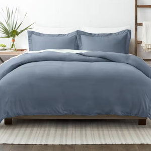 A blue king-size duvet cover set with two matching pillow shams, featuring a smooth and solid design. The bed is styled with a clean, modern look, complemented by a white area rug and a wooden side table with a small plant.