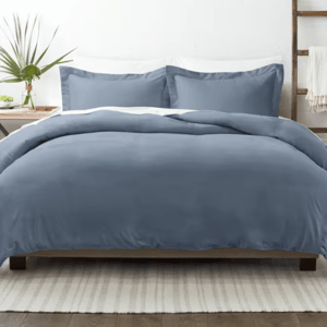 A blue king-size duvet cover set with two matching pillow shams, featuring a smooth and solid design. The bed is styled with a clean, modern look, complemented by a white area rug and a wooden side table with a small plant.