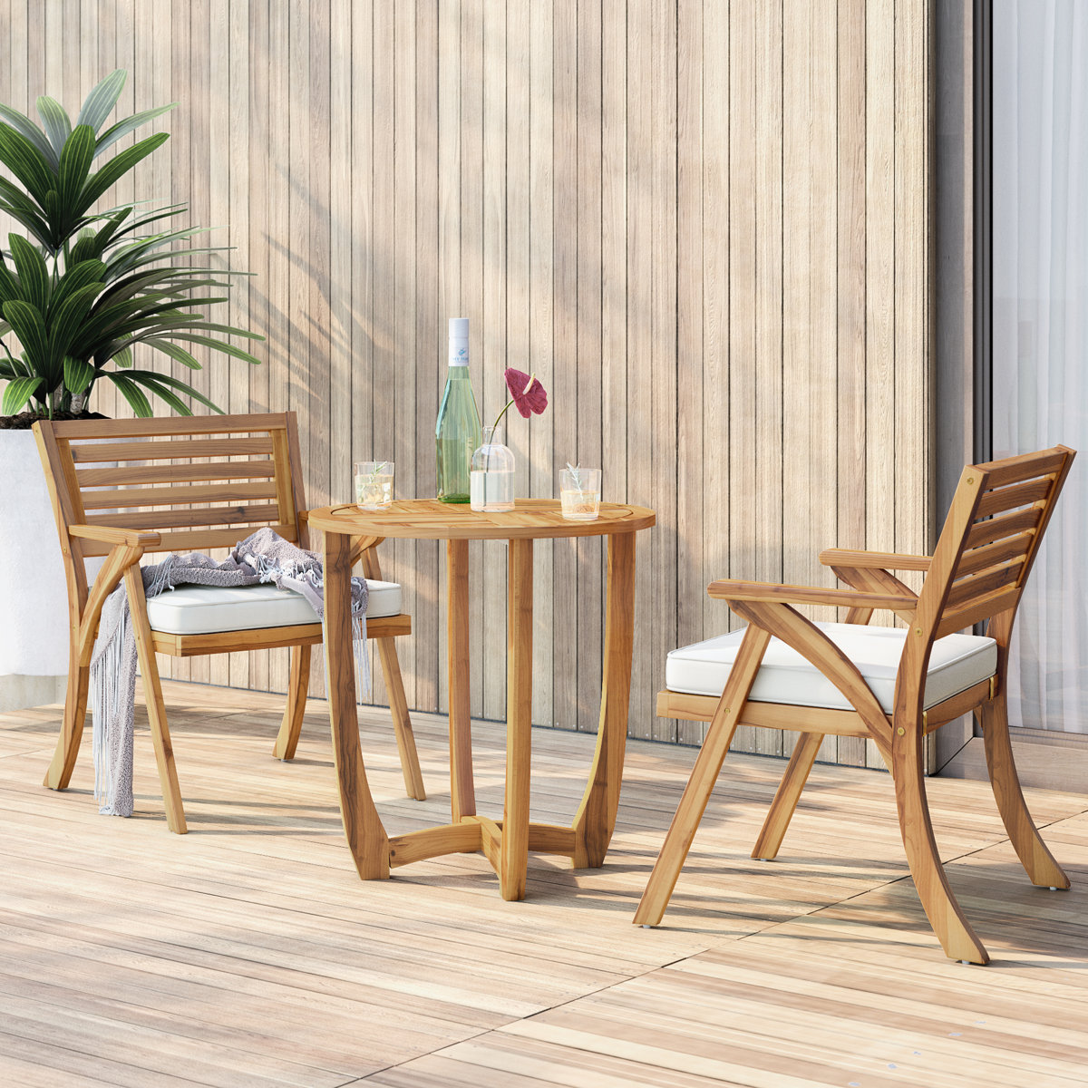 A solid wood patio set includes a round table and two chairs with slatted backs and armrests. The chairs have white cushions, and a small plant and drinks are on the table, set against a wood-paneled wall.