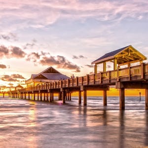 A wooden pier extends over the ocean at sunset, with two covered pavilions and a vibrant sky of orange and purple hues reflecting on the water below.