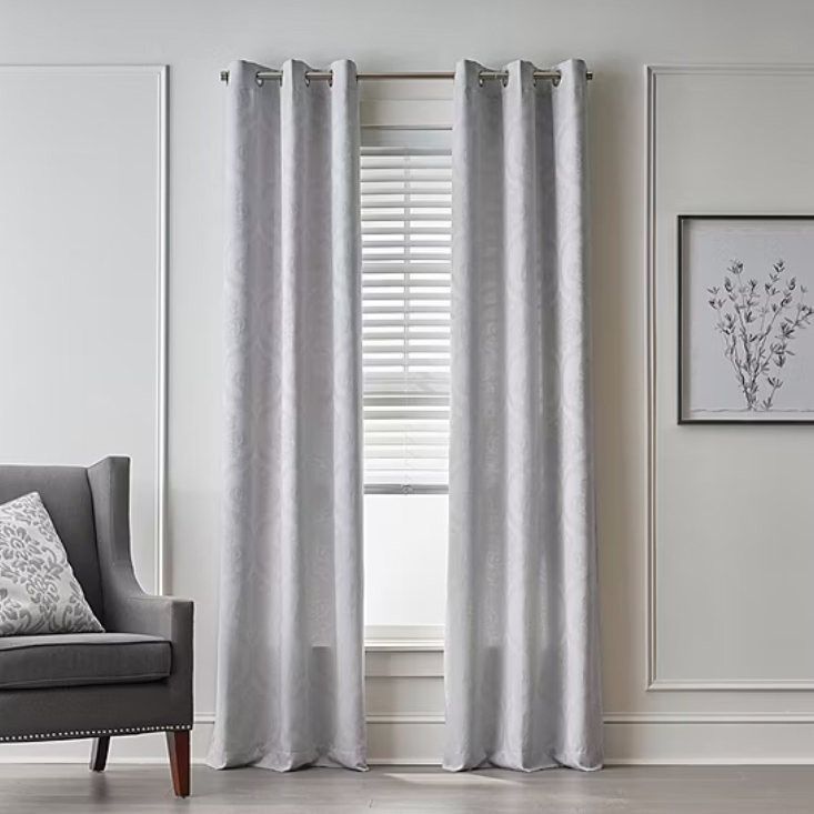 Light-Filtering Jacquard Curtains in a soft, neutral color hang elegantly with grommets on a curtain rod. They frame a window with blinds and are complemented by a gray armchair and a botanical wall print, creating a calming and sophisticated atmosphere.