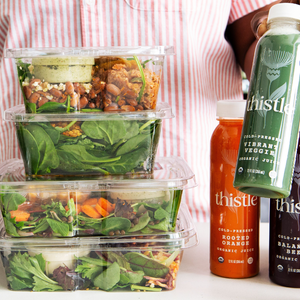 Stack of salad containers and bottles of Thistle cold-pressed organic juices, including \“Vibrant Veggie,\“ \“Rooted Orange,\“ and \“Balanced Beet.\“