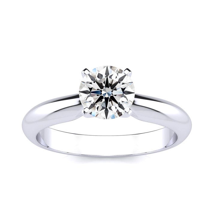 A solitaire ring featuring a 1-carat round diamond set in a simple, polished metal band with a classic four-prong setting. The band has a smooth, reflective finish, enhancing the diamond's brilliance.