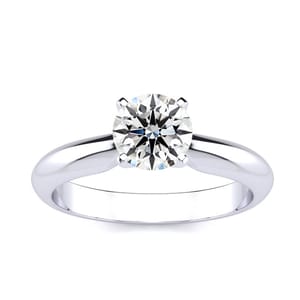 A solitaire ring featuring a 1-carat round diamond set in a simple, polished metal band with a classic four-prong setting. The band has a smooth, reflective finish, enhancing the diamond's brilliance.