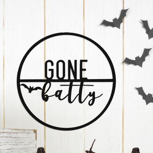A round black wall sign with the text \“Gone Batty,\“ featuring a bat design. Surrounding it are additional small bat cutouts on a white wooden background.