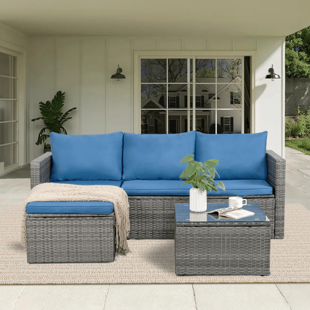 Outdoor patio set with gray wicker frame, blue cushions, a matching ottoman, and a small glass-topped coffee table. Includes a throw blanket and a white vase with a plant, all set on a light-colored rug under a covered porch.