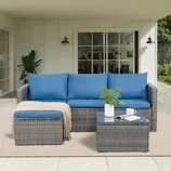 Outdoor patio set with gray wicker frame, blue cushions, a matching ottoman, and a small glass-topped coffee table. Includes a throw blanket and a white vase with a plant, all set on a light-colored rug under a covered porch.