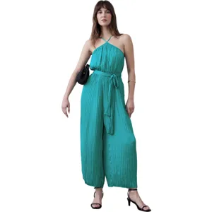 A woman models a turquoise, pleated halter-neck jumpsuit, paired with black strappy high heels and a black shoulder bag.