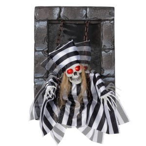 Hanging skeleton decoration with red glowing eyes, dressed in a black and white striped outfit and hat, set in a stone-like frame with chains.
