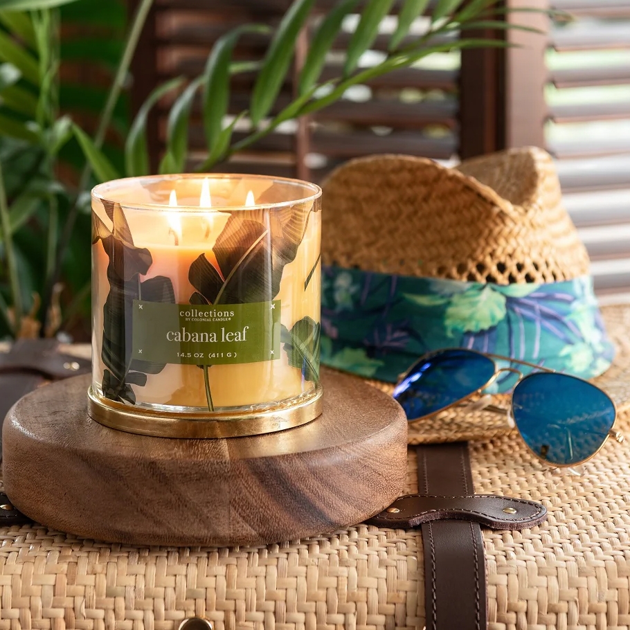 A \“Cabana Leaf\“ candle sits on a wooden base, next to a straw hat with a floral band and blue sunglasses, all atop a woven surface.