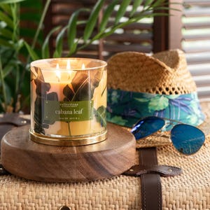 A \“Cabana Leaf\“ candle sits on a wooden base, next to a straw hat with a floral band and blue sunglasses, all atop a woven surface.