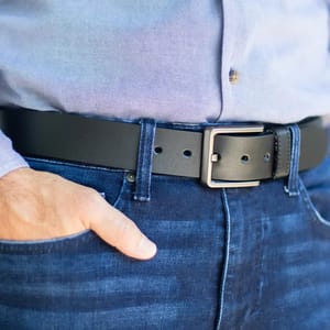 A person wearing blue jeans and a light blue shirt with a black leather belt featuring a rectangular silver buckle.