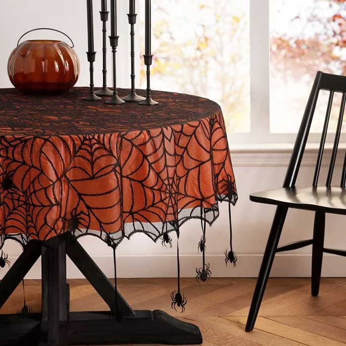 A black and orange spider web tablecloth with hanging spider decorations adorns a round table, accompanied by a black chair and a tall candle holder.