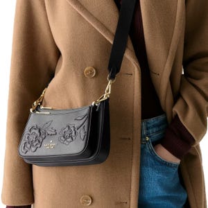 A person is wearing a tan coat and blue jeans, carrying a black Kate Spade shoulder bag with floral embroidery and gold hardware.