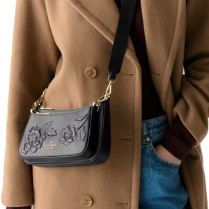 A person is wearing a tan coat and blue jeans, carrying a black Kate Spade shoulder bag with floral embroidery and gold hardware.