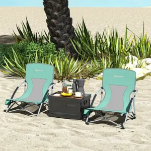 Two teal and gray beach chairs are placed on sand next to a palm tree, accompanied by a small black table with drinks and snacks.