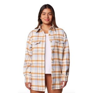 A woman is wearing a check-patterned flannel shirt-jacket in shades of beige, light blue, and white over a white top and tan pants. The jacket features button closures and chest pockets.