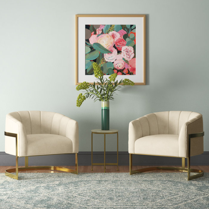 Two beige armchairs with gold accents flank a small round table with a teal vase and greenery, set against a pale blue wall with a framed floral painting. A textured blue-gray rug covers the floor.