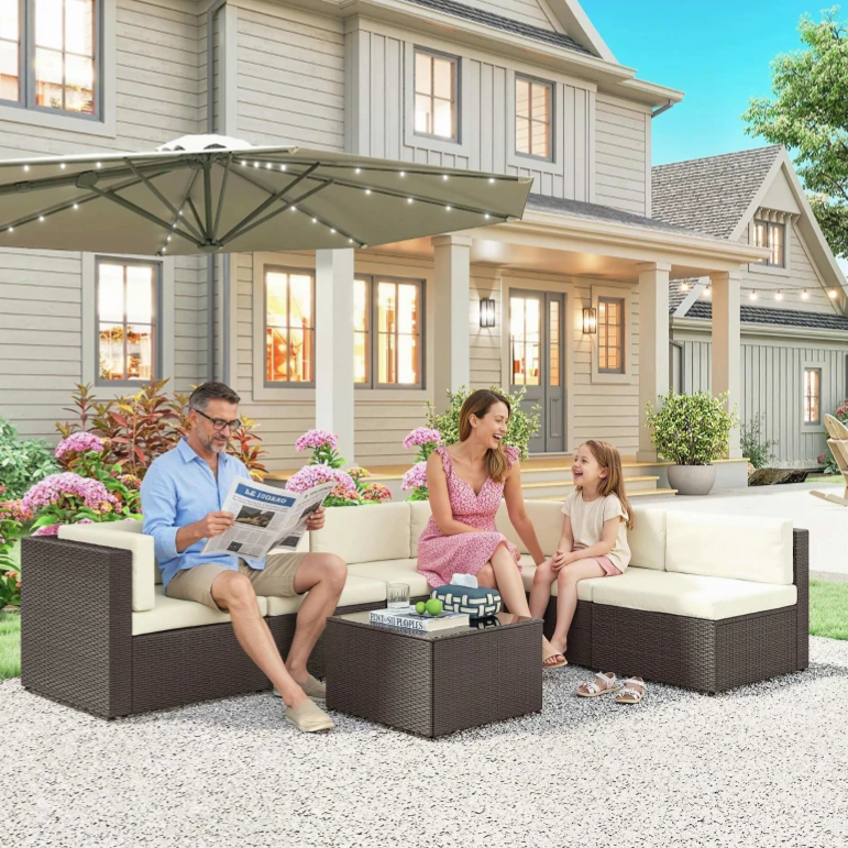 A family is relaxing on a wicker outdoor patio set with beige cushions, under a large umbrella with string lights. The setting includes a modern house backdrop with decorative plants and flowers.