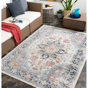 A 5' x 7' area rug with an intricate pattern of blue, orange, and beige hues is placed on a tiled floor. It complements outdoor wicker furniture, which features white cushions, a red throw, and decorative items like a blue ceramic pitcher and potted plants.