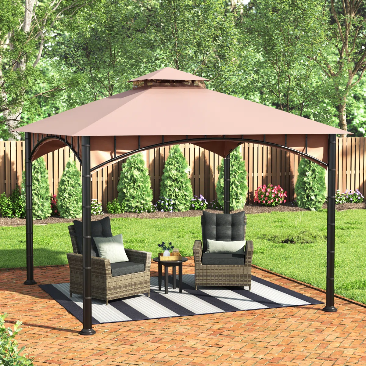 A backyard gazebo with a peaked roof, featuring two wicker armchairs with cushions, a small round table on a striped rug, set on a brick patio with a wooden fence and greenery in the background.