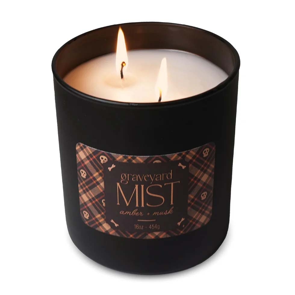 A lit candle in a black container labeled \“Graveyard Mist\“ with scents of amber and musk, weighing 16oz (454g). The label has a plaid design with skull and bone motifs.