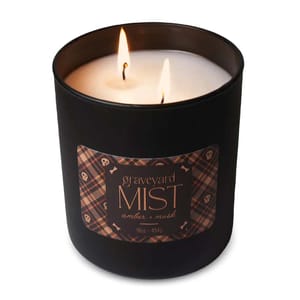 A lit candle in a black container labeled \“Graveyard Mist\“ with scents of amber and musk, weighing 16oz (454g). The label has a plaid design with skull and bone motifs.