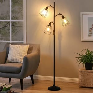A modern 70\“ floor lamp with three exposed bulbs and black metal frames, featuring a sleek vertical design. It stands beside a gray armchair, with a woven side table and a potted plant nearby, casting warm light onto the room.
