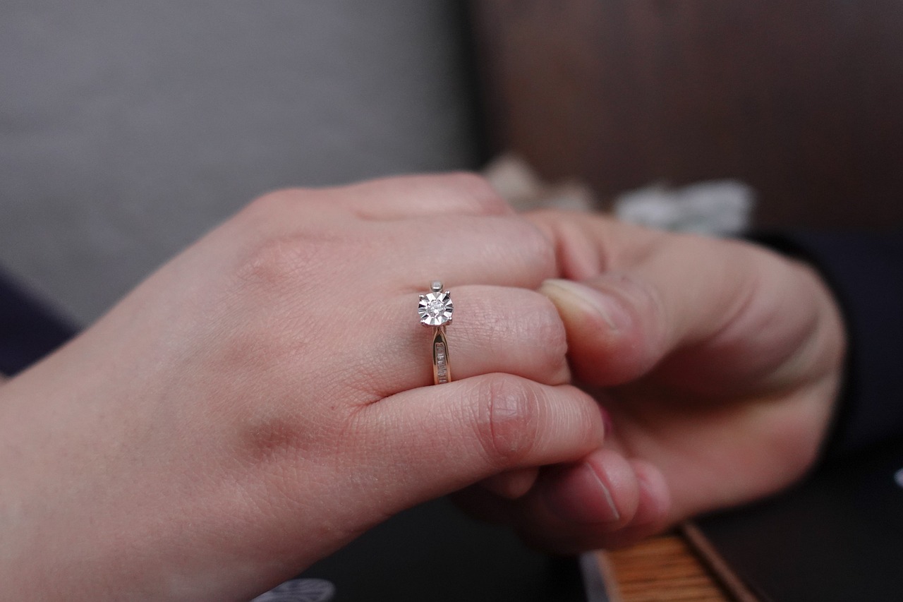 A hand wearing a diamond solitaire ring on the ring finger.