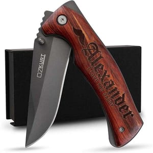 A folding knife featuring a wooden handle engraved with \“Alexander I Love You Daddy\“ and accompanied by a black gift box. The blade is branded \“OZKURT.\“
