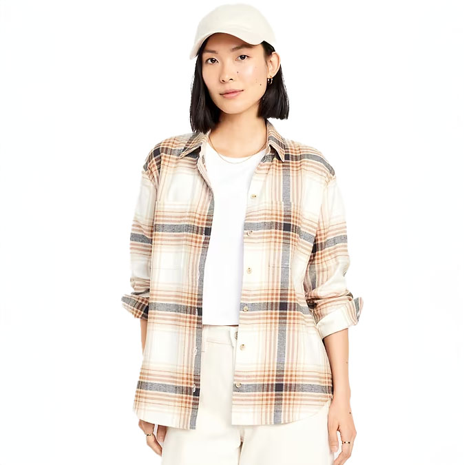 A woman is wearing a beige plaid oversized boyfriend flannel shirt with a white t-shirt, beige pants, and a white baseball cap.