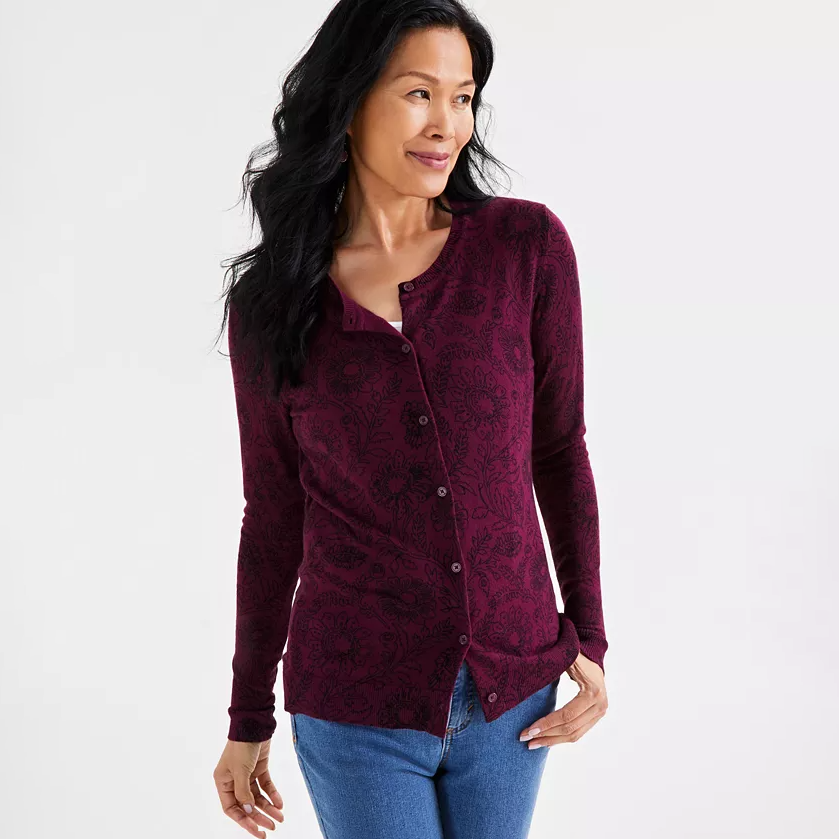 A woman is wearing a long-sleeve, burgundy cardigan with a subtle floral print, buttoned front, and a rounded neckline, paired with blue jeans.