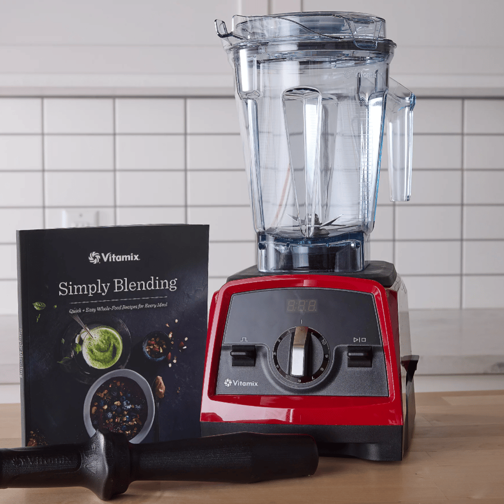 A Vitamix blender with a red base, clear pitcher, and accompanying tamper is displayed alongside a Vitamix cookbook titled \“Simply Blending\“ on a kitchen counter with a tiled backsplash.