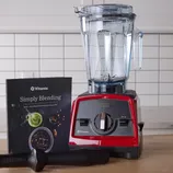 A Vitamix blender with a red base, clear pitcher, and accompanying tamper is displayed alongside a Vitamix cookbook titled \“Simply Blending\“ on a kitchen counter with a tiled backsplash.