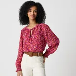 Woman wearing a vibrant floral blouse in shades of pink and orange, paired with white pants and a brown belt.