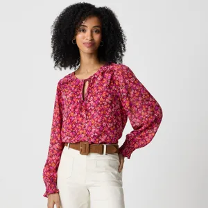 Woman wearing a vibrant floral blouse in shades of pink and orange, paired with white pants and a brown belt.