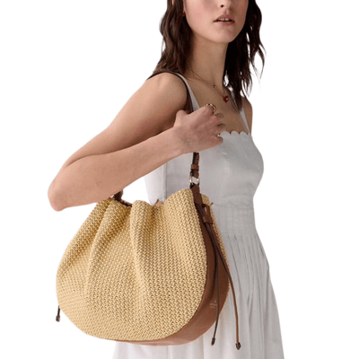 A woman in a white dress holds a large woven tote bag with leather straps.
