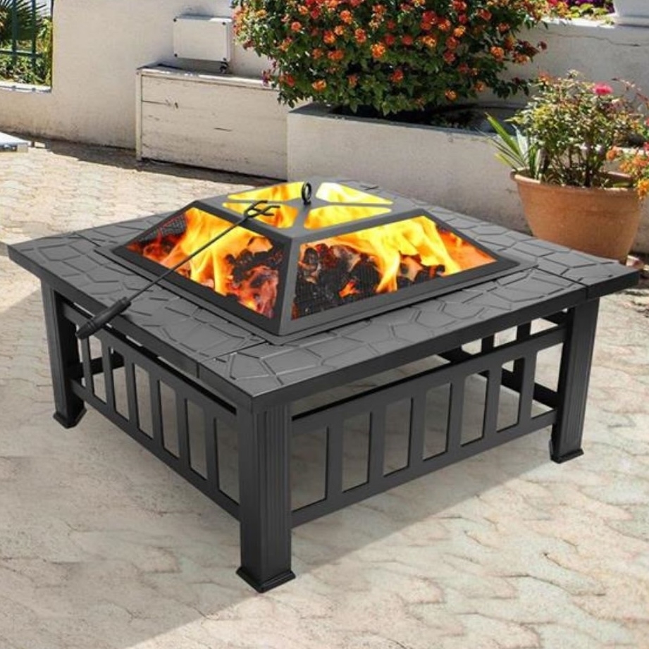 A black metal fire pit with a mesh cover, featuring a square design and accompanied by a fire poker, situated on a patio with surrounding plants.