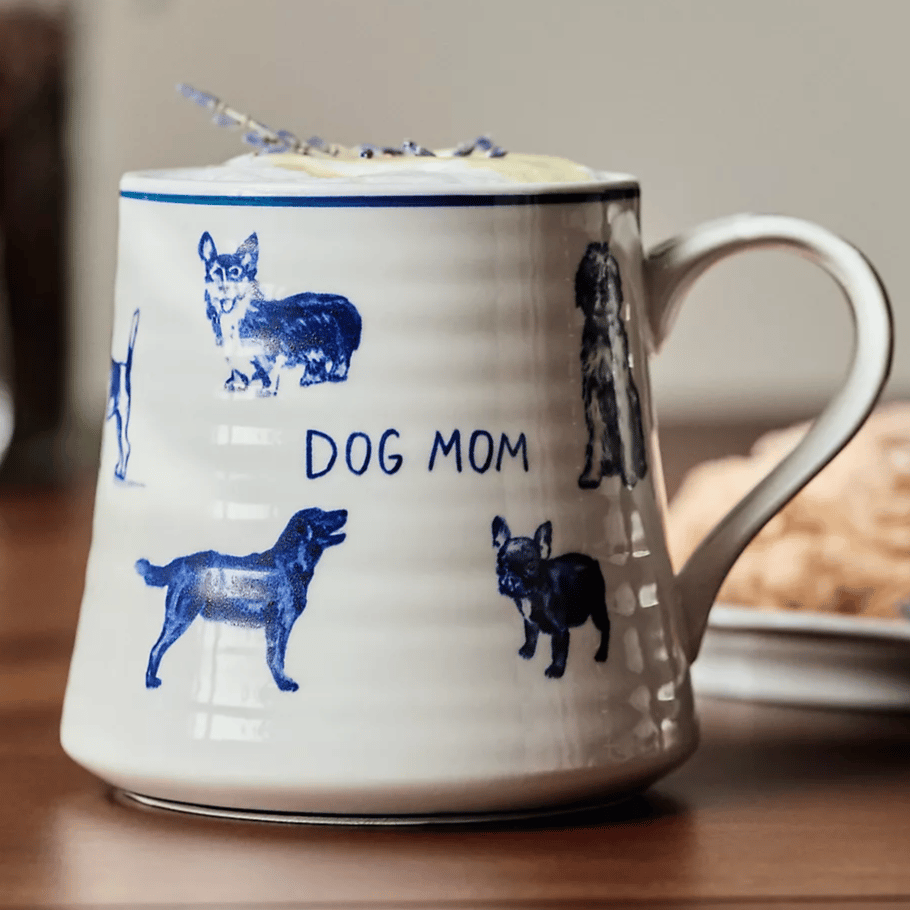 A ceramic mug with the phrase \“Dog Mom\“ features blue illustrations of various dog breeds and has a lavender garnish on the rim.