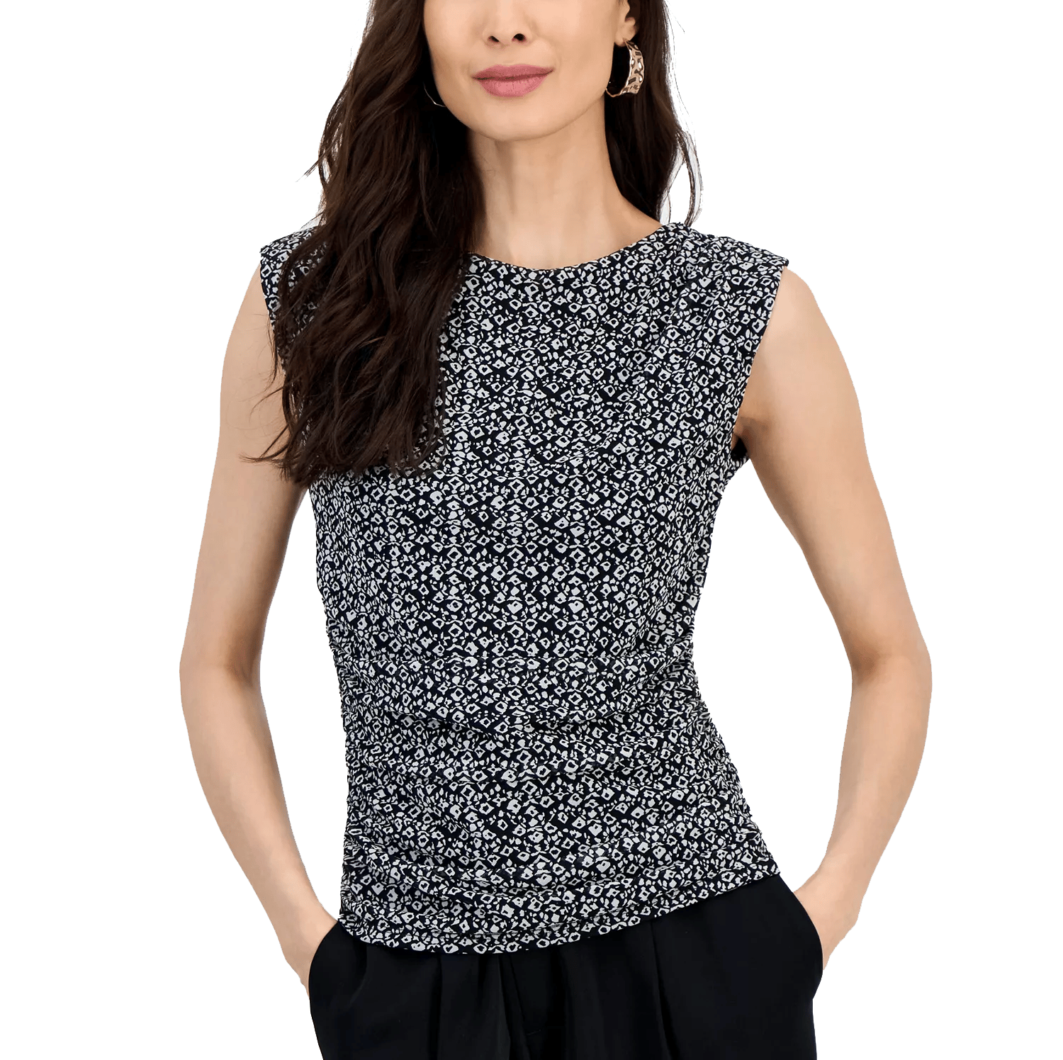 A woman is wearing a sleeveless, black and white patterned top with a fitted design, paired with black trousers. She has long dark hair and is accessorizing with hoop earrings, standing with hands in her pockets.