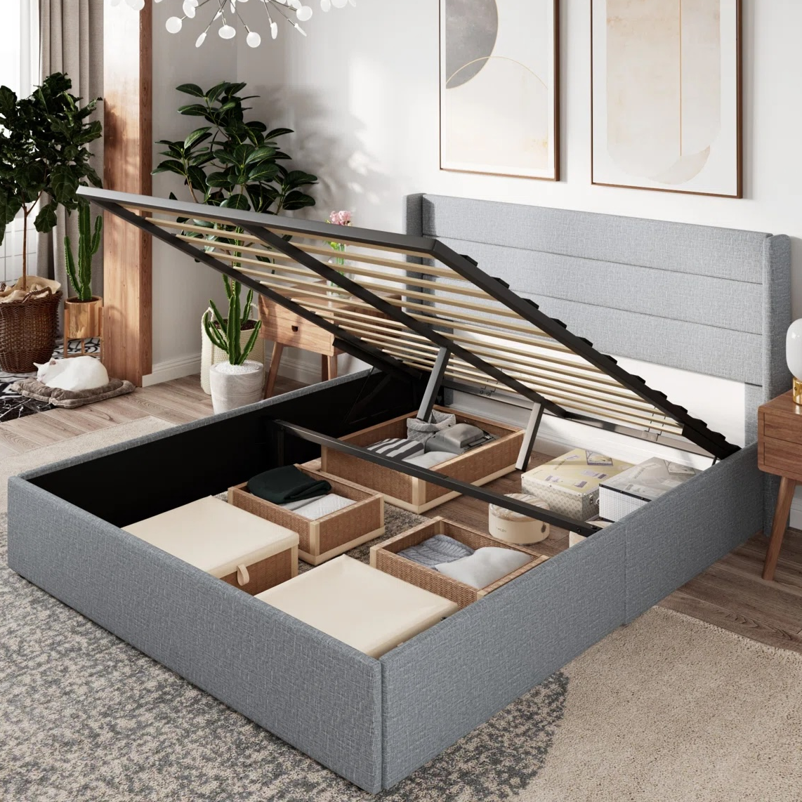 A gray upholstered storage bed with a lift-up platform reveals organized compartments filled with storage bins. The room features modern decor, including framed artwork, potted plants, and wooden furniture.