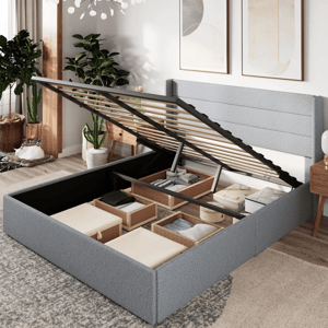 A gray upholstered storage bed with a lift-up platform reveals organized compartments filled with storage bins. The room features modern decor, including framed artwork, potted plants, and wooden furniture.
