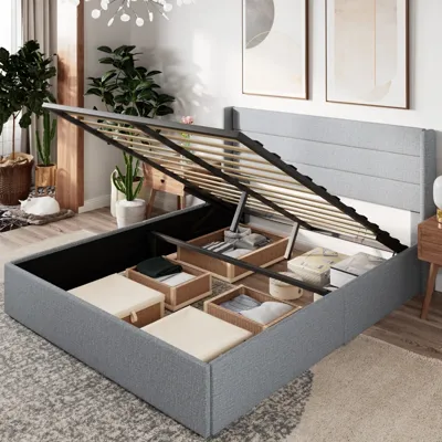 A gray upholstered storage bed with a lift-up platform reveals organized compartments filled with storage bins. The room features modern decor, including framed artwork, potted plants, and wooden furniture.