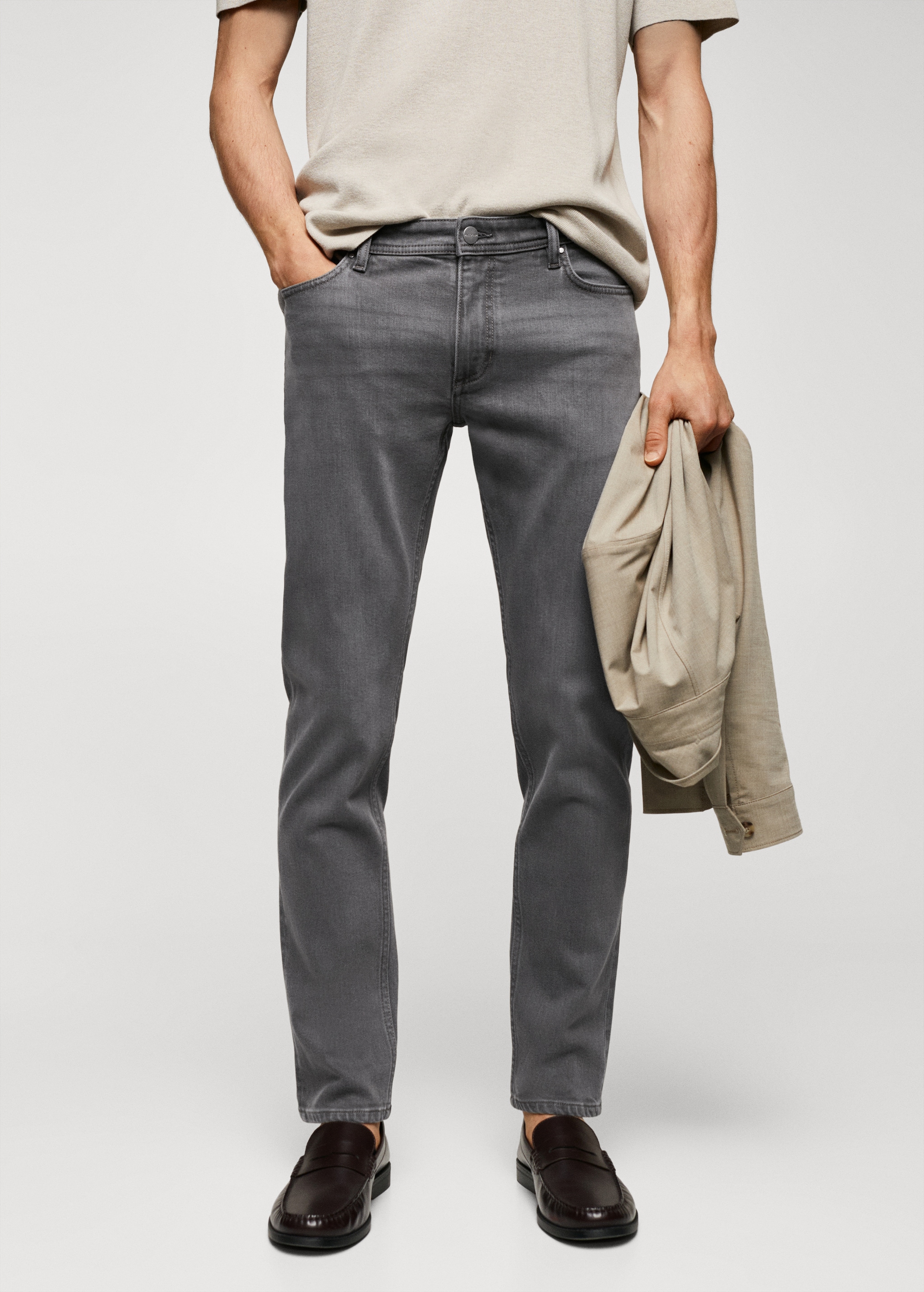 Gray slim-fit jeans with a standard waist and five pockets, paired with dark brown loafers and a beige jacket being held.