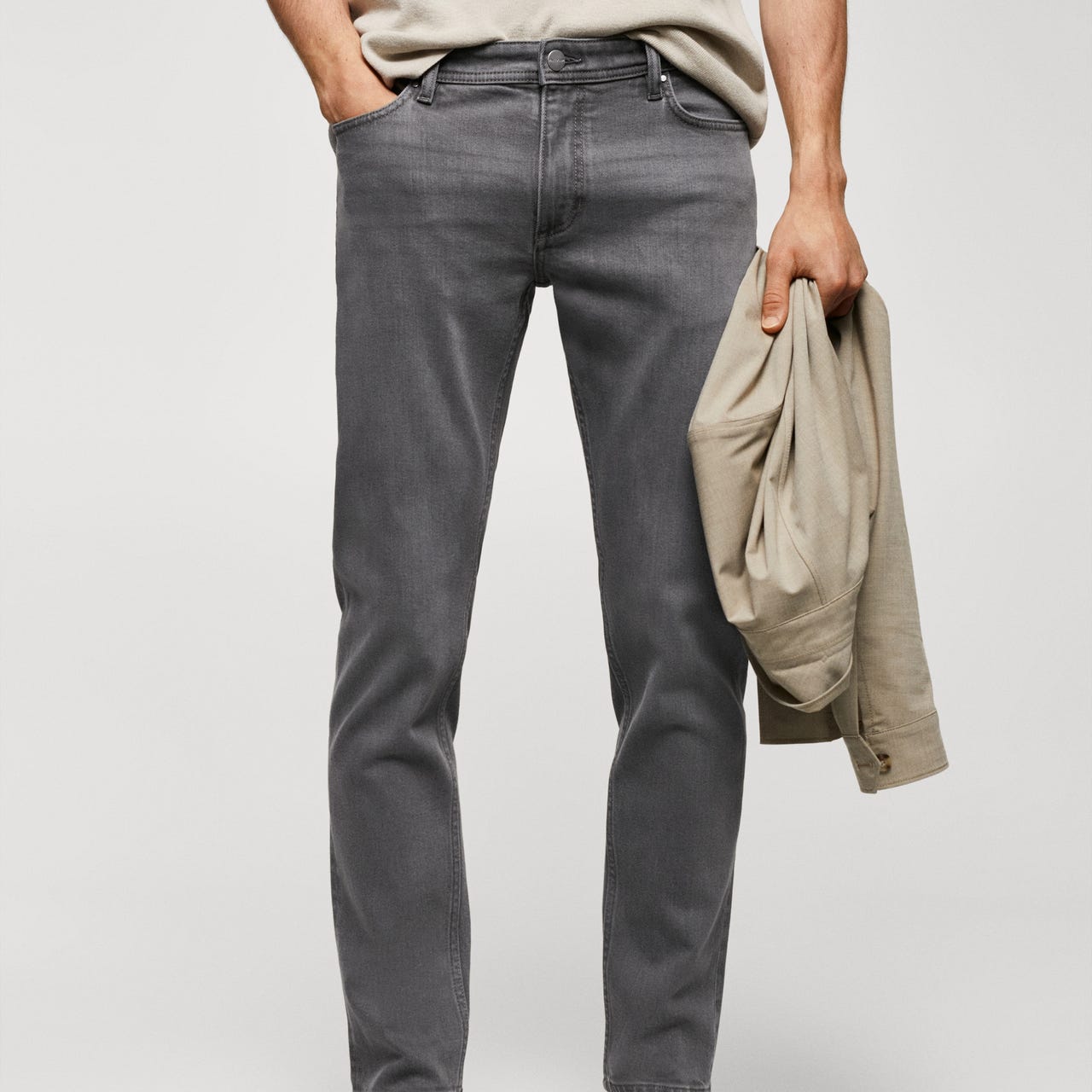 Gray slim-fit jeans with a standard waist and five pockets, paired with dark brown loafers and a beige jacket being held.