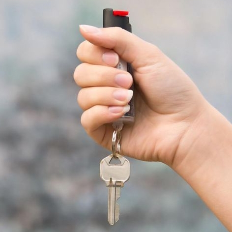 A hand holds a keychain pepper spray with a red button, attached to a single metal key.