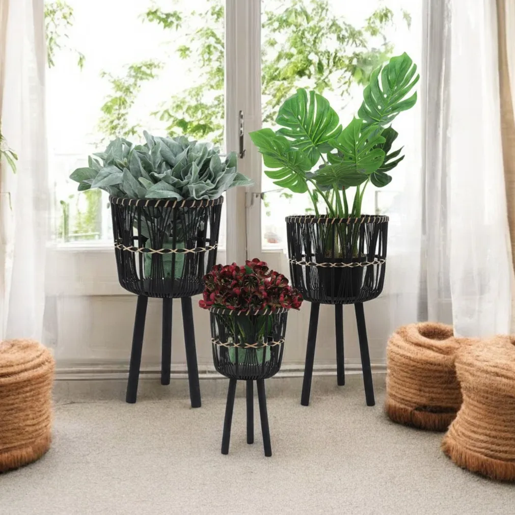 Three black woven planters with tripod legs, featuring different plants, are displayed indoors. They are set against a white curtain backdrop with two cylindrical woven baskets nearby.