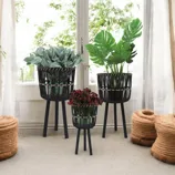Three black woven planters with tripod legs, featuring different plants, are displayed indoors. They are set against a white curtain backdrop with two cylindrical woven baskets nearby.