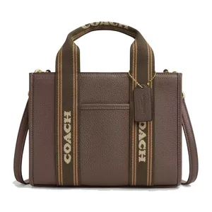 A brown Coach tote bag with textured leather, featuring dual fabric handles adorned with the Coach logo, a detachable shoulder strap, and a branded tag hanging from a gold chain.