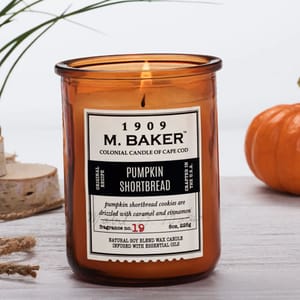 An amber jar candle labeled \“1909 M. Baker\“ with a pumpkin shortbread scent, featuring notes of caramel and cinnamon. Candle is an 8 oz natural soy blend wax infused with essential oils.